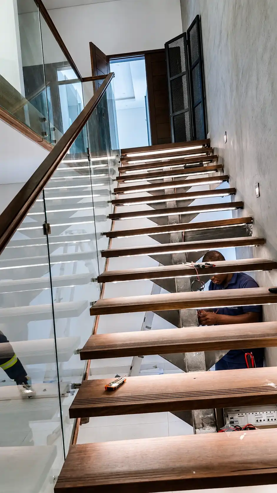 Technician wiring LED profile light strips under a floating wooden staircase in Hoima